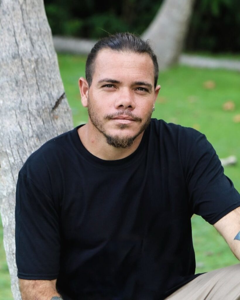 Portrait of Tony Fuertes in a black T-shirt, seated beside a palm trunk